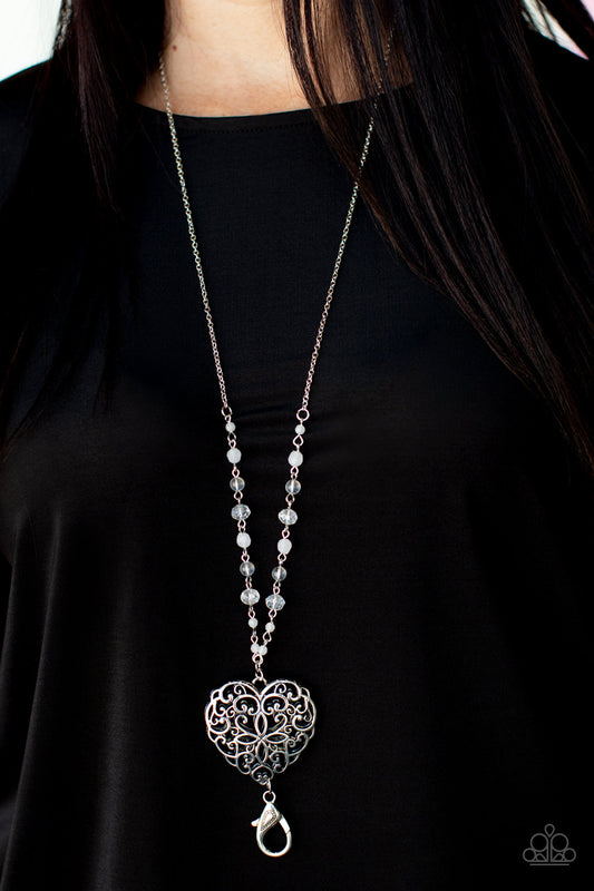 A dainty collection of opaque, glassy, and crystal-like beads give way to an oversized filigree filled heart frame, creating an antiqued locket inspired pendant at the bottom of a lengthened silver chain. A lobster clasp hangs from the bottom of the design to allow a name badge or other item to be attached. Features an adjustable clasp closure.  Sold as one individual lanyard. Includes one pair of matching earrings.