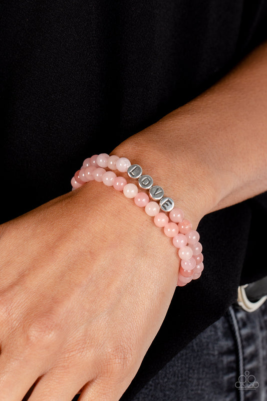 Cloudy beads in various shades of pink extend around the wrist on elastic stretchy bands for a two-for-one romantic stack. Centered on one of the colorful bracelets, silver discs stamped with individual black letters spell out the word "love."  Sold as one set of two bracelets.