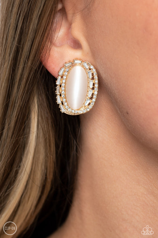 Rows of white rhinestones, gold studs, and white gem bars create dramatic dimension encircling an elongated white cat's eye stone for a sparkly statement piece. Earring attaches to a standard clip-on fitting.  Sold as one pair of clip-on earrings.  Clip On Earring