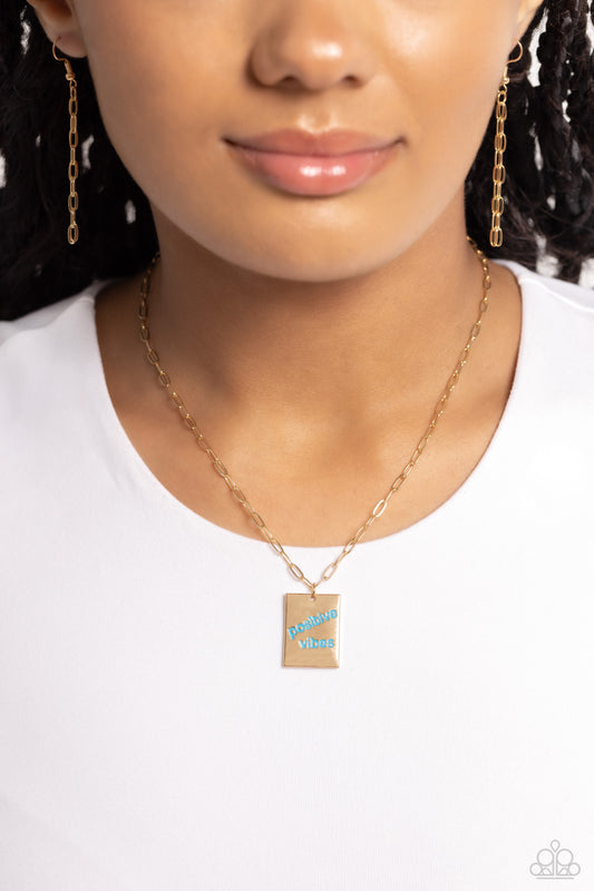 A rectangular gold pendant is stamped with the phrase "positive vibes" in a vibrant blue font at the bottom of a trendy, dainty gold paperclip chain, creating an inspiring pendant below the collar. Features an adjustable clasp closure.  Sold as one individual necklace. Includes one pair of matching earrings.
